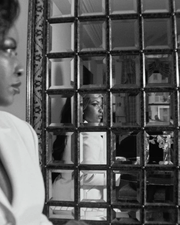 Monochrome portrait of an African-American woman in white formalwear, partially reflected in a grid-patterned decorative mirror.