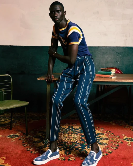 An African American adult male in an indoor fashion shot, modeling a blue t-shirt and striped pants, and matching blue shoes, leaning on a table.