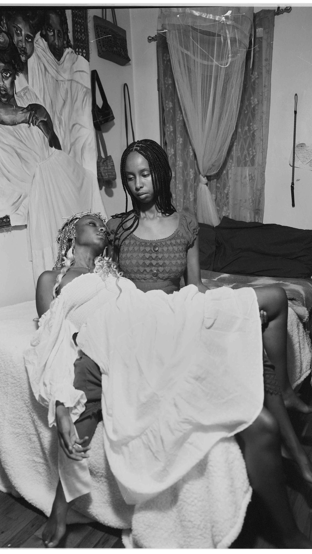 Black-and-white image features two women indoors, one lying across the lap of the other, with a mural visible in the background.