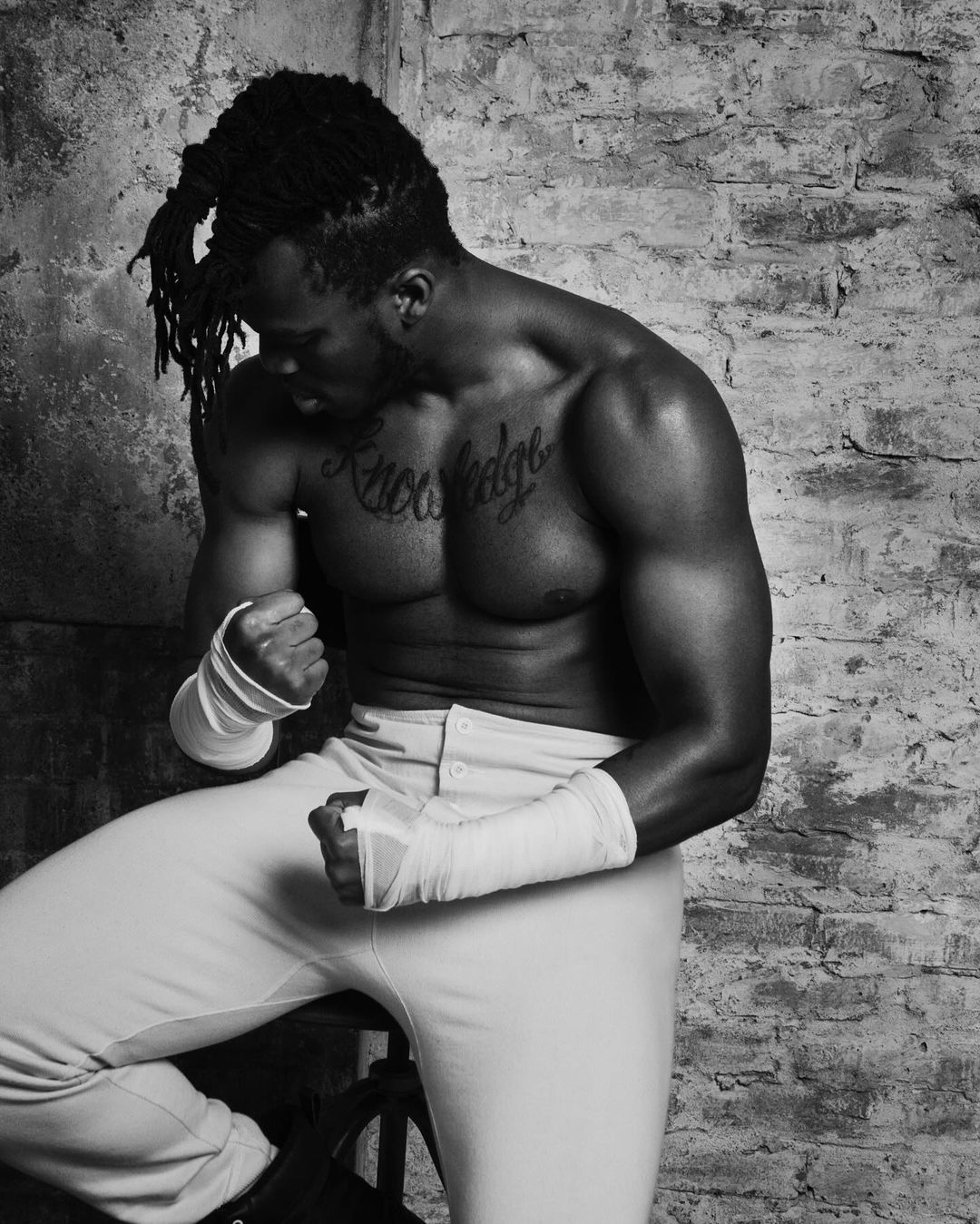 Black and white shot of a muscular African American man with dreadlocks sitting shirtless against a brick wall with wrapped hands.