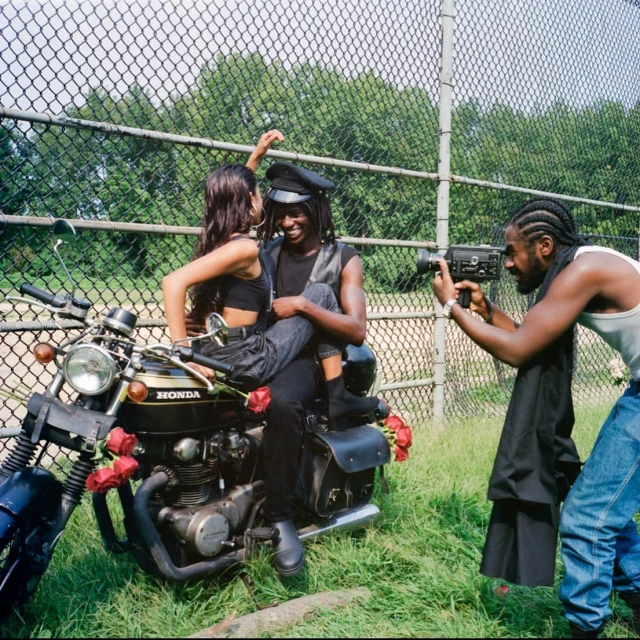 Young couple poses on a vintage motorcycle decorated with red roses, while a man films them in an outdoor grassy setting.