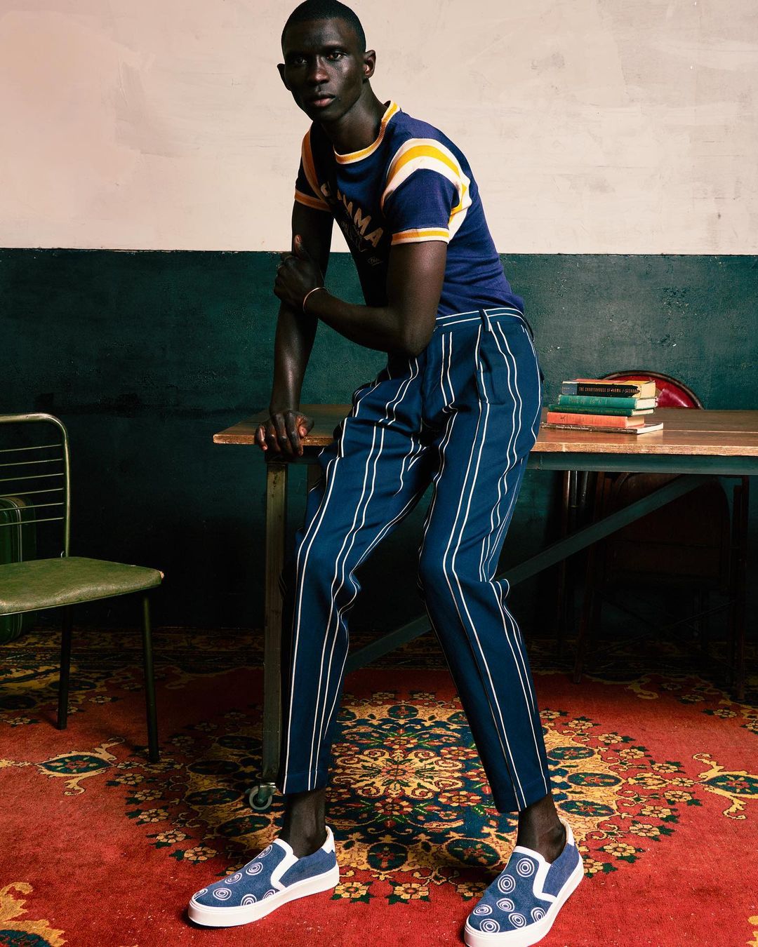 A man poses in a blue t-shirt with yellow stripes and navy pants with white stripes in an indoor setting, on a patterned rug.
