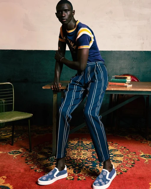 A man poses in a blue t-shirt with yellow stripes and navy pants with white stripes in an indoor setting, on a patterned rug.