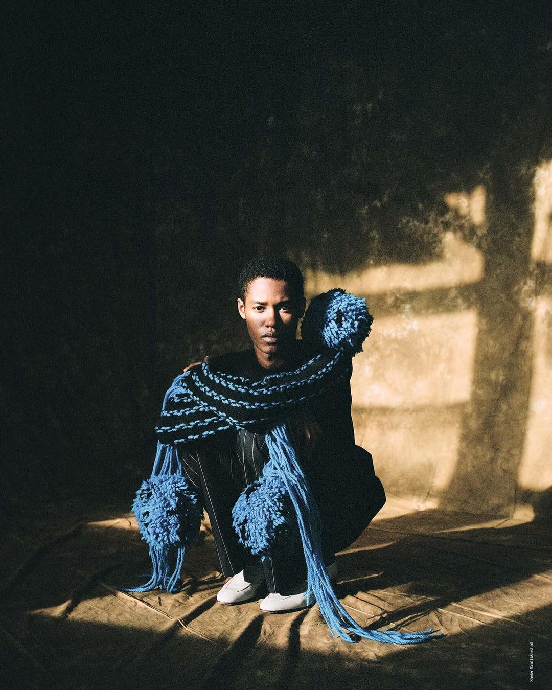 Portrait of an androgynous person with short hair squatting on the ground, adorned with blue yarn, in a spotlight in a dark space.