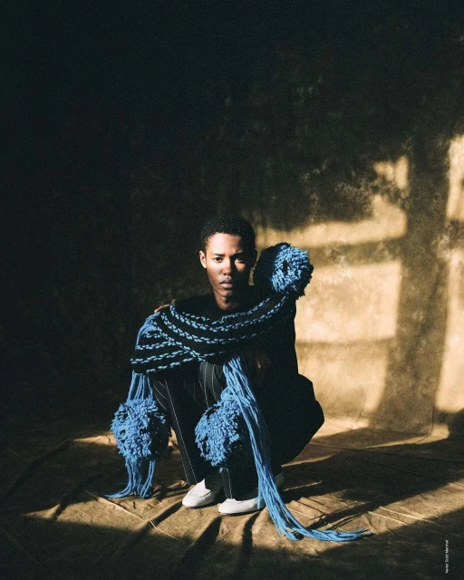 Portrait of an androgynous person with short hair squatting on the ground, adorned with blue yarn, in a spotlight in a dark space.