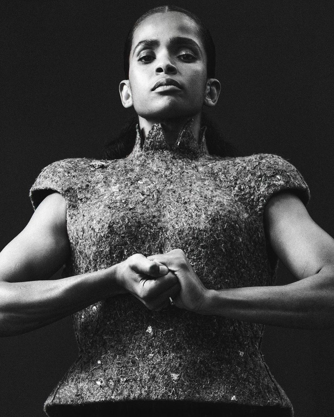 Monochrome portrait of adult woman with hands clasped, wearing a textured, high-necked top against a dark backdrop in a studio setting.