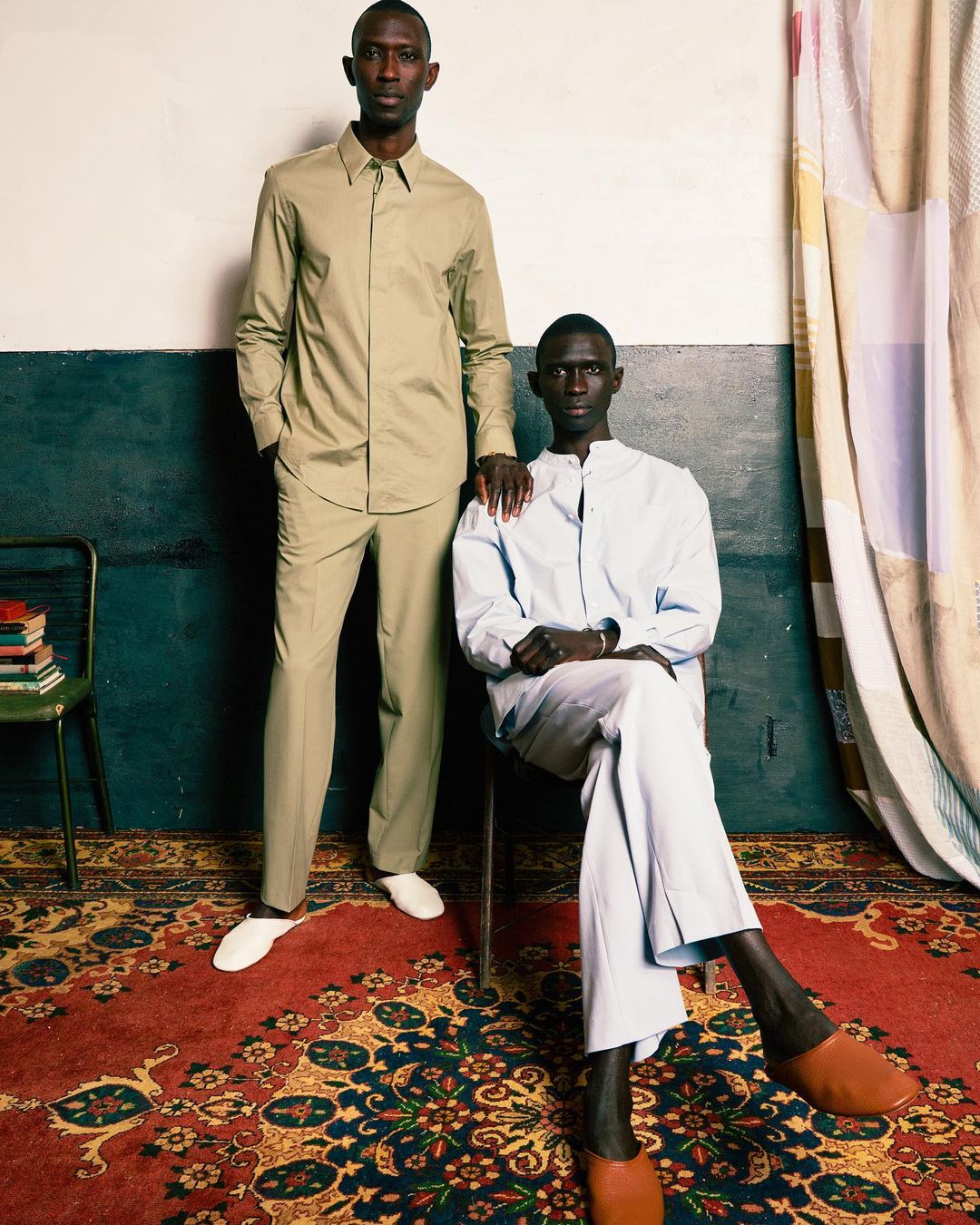 Two dark-skinned men in stylish, light-colored shirts and pants pose in an indoor setting, showcasing fashion and design with a patterned carpet.