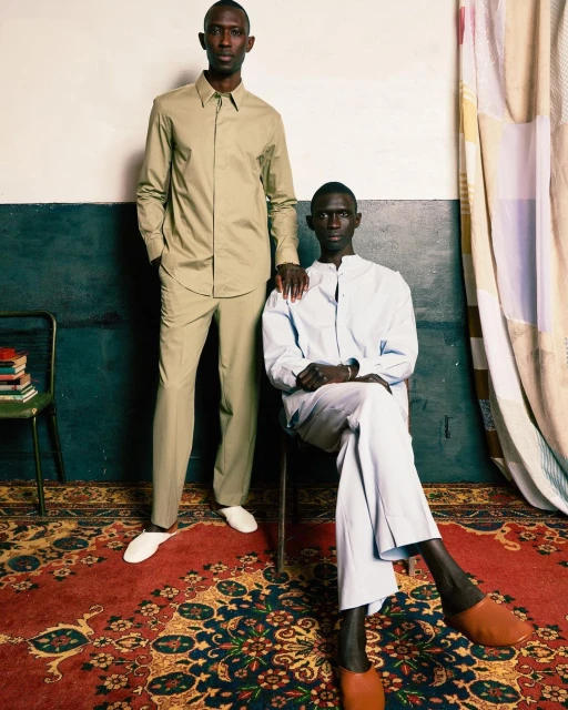 Two dark-skinned men in stylish, light-colored shirts and pants pose in an indoor setting, showcasing fashion and design with a patterned carpet.