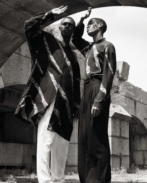 Two Black men pose beneath a stone archway. One wears a long, striped jacket with white pants, and the other wears a striped top with black trousers.