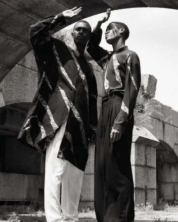Two Black men pose beneath a stone archway. One wears a long, striped jacket with white pants, and the other wears a striped top with black trousers.