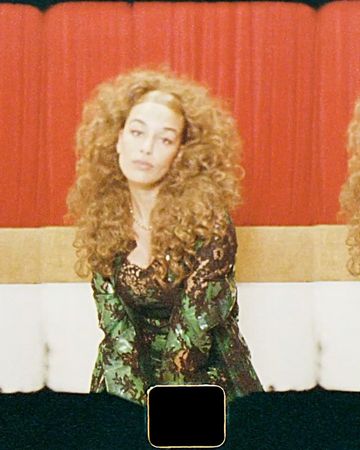 Woman with curly hair in green floral dress poses on a white couch in front of a red curtain in an eye-level medium shot.