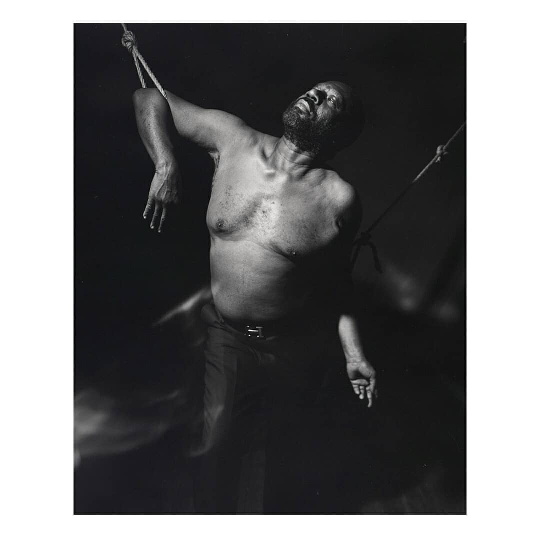 Monochrome portrait of an African American man suspended by ropes in a dimly lit space, head tilted back, eyes closed, and shirtless.