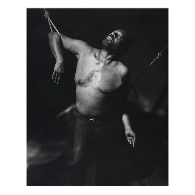 Monochrome portrait of an African American man suspended by ropes in a dimly lit space, head tilted back, eyes closed, and shirtless.