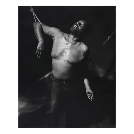 Monochrome portrait of an African American man suspended by ropes in a dimly lit space, head tilted back, eyes closed, and shirtless.