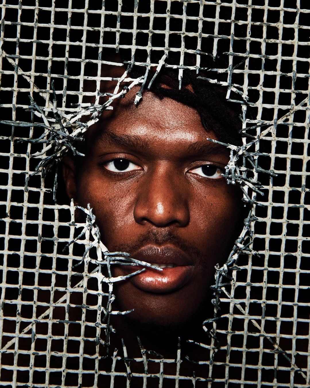 Close-up studio portrait of a Black adult male with dreadlocks, lips slightly parted, framed by a white lattice with barbed wire details.