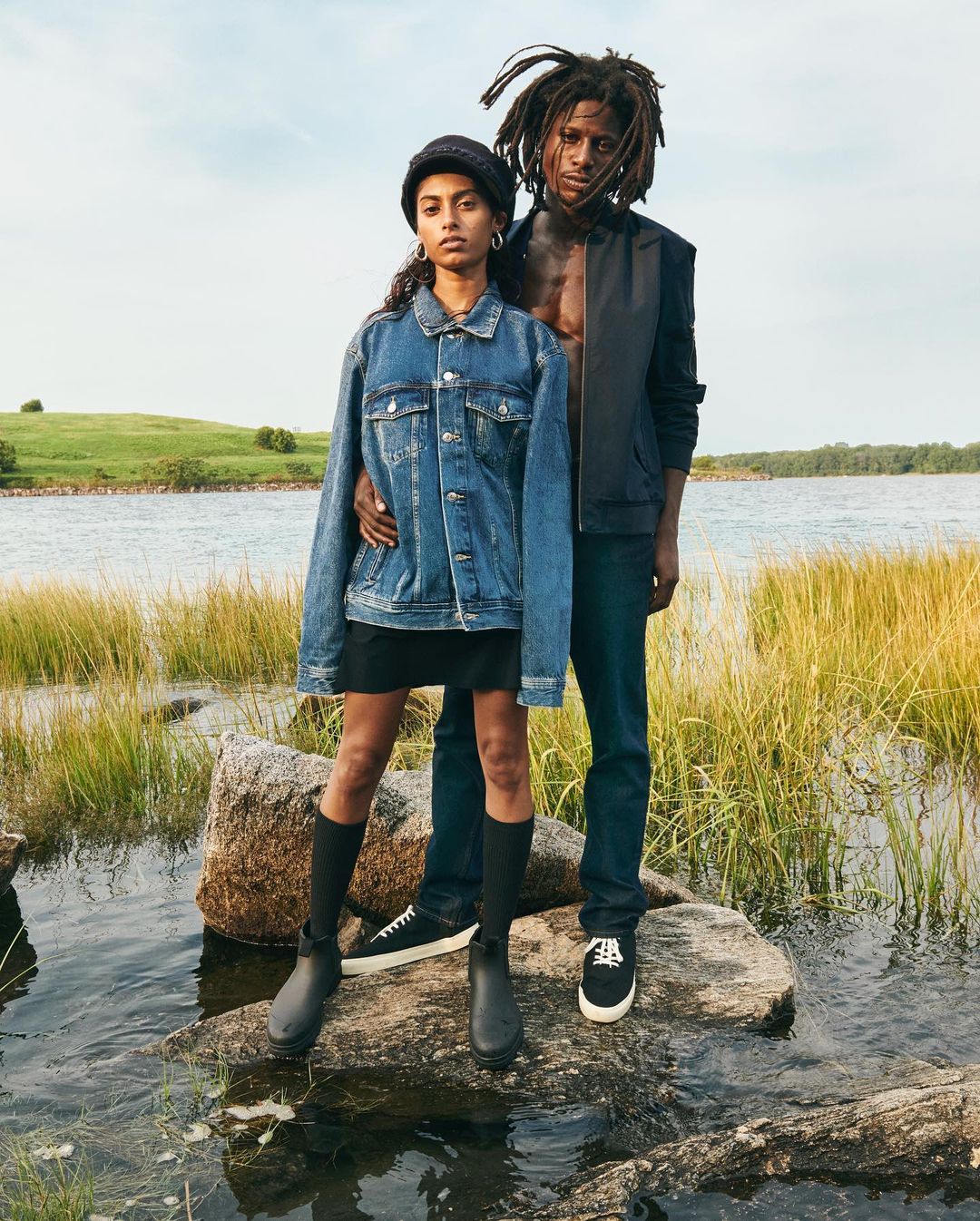 A young couple stands on rocks in a marshy field by a lake. The woman wears a denim jacket and the man a black vest.