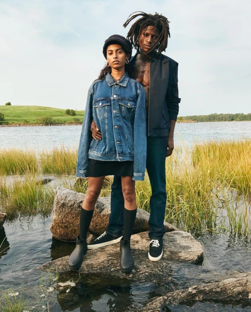 A young couple stands on rocks in a marshy field by a lake. The woman wears a denim jacket and the man a black vest.