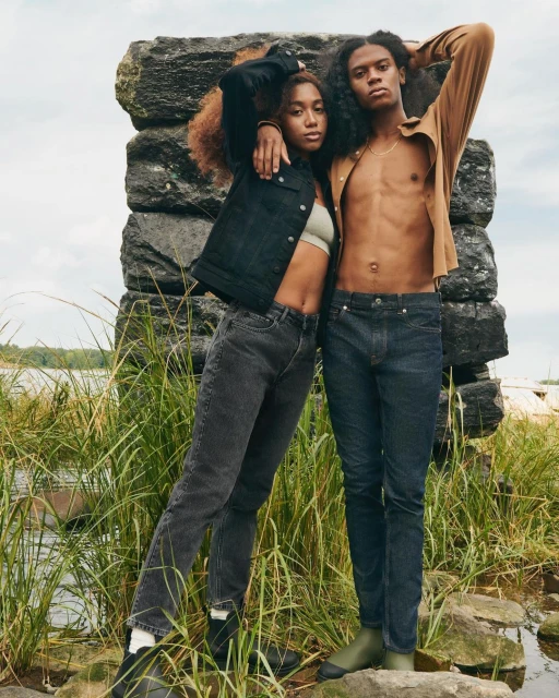 A fashionable couple poses outdoors in front of a stone structure, wearing jeans and jackets with natural light highlighting their features.