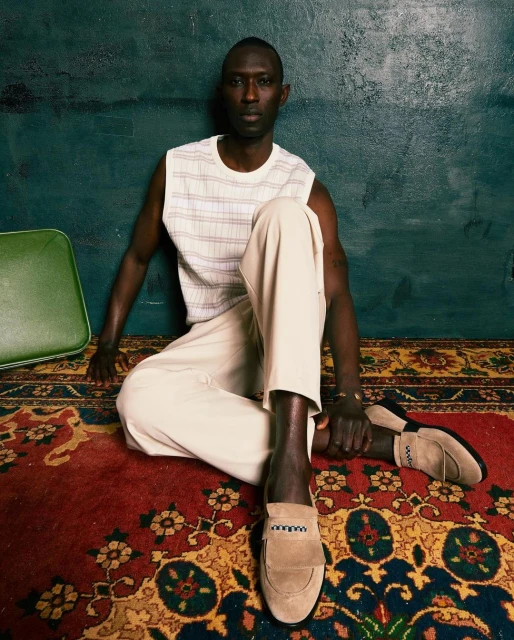 Adult male poses indoors wearing a striped tank top, white pants, and loafers, sitting on a red patterned rug against a teal wall.