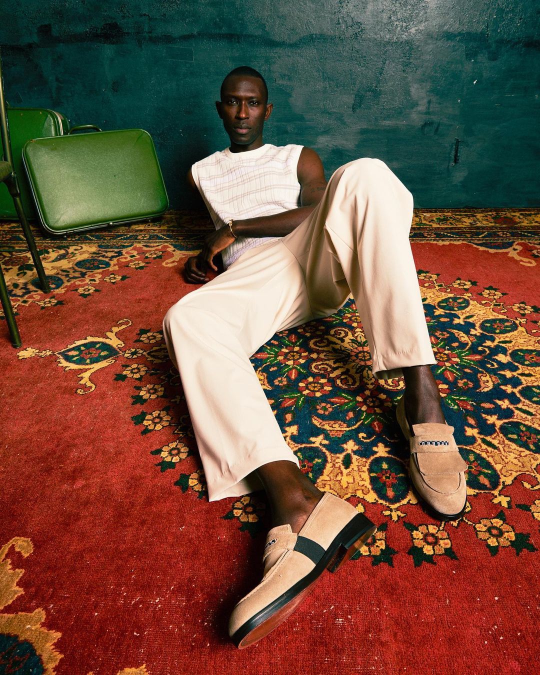 A slender Black man lounges on a red patterned rug in cream pants and knit shirt near green vintage luggage, looking at the camera.