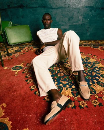 A slender Black man lounges on a red patterned rug in cream pants and knit shirt near green vintage luggage, looking at the camera.