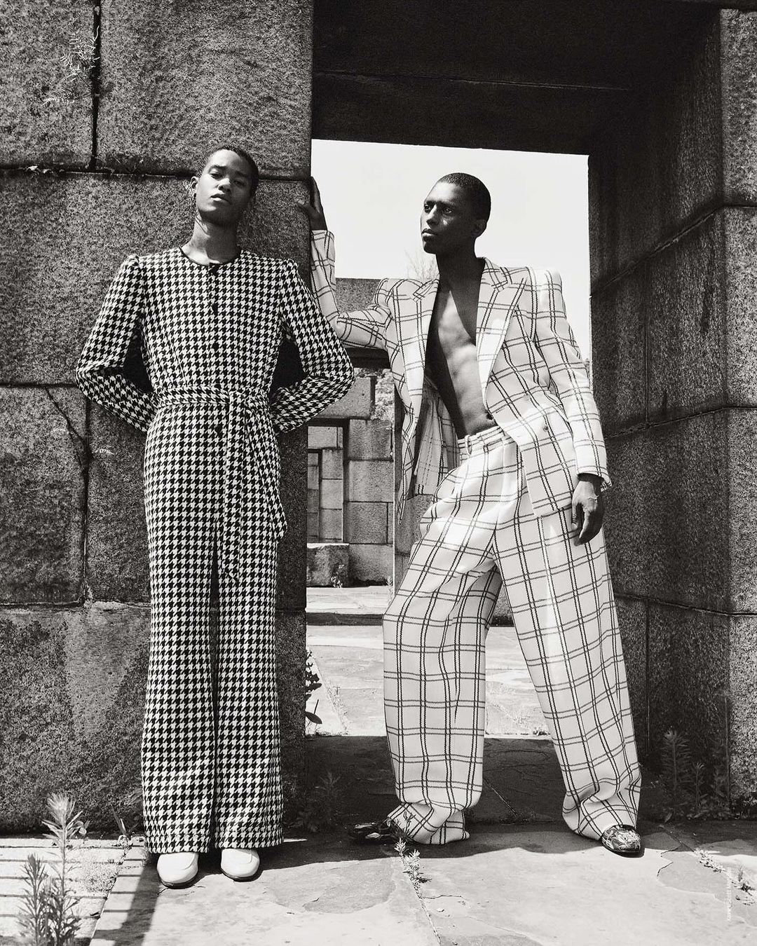 Two Black male fashion models in patterned suits pose near a stone archway, showcasing a black and white editorial fashion aesthetic.