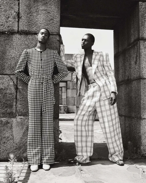 Two Black male fashion models in patterned suits pose near a stone archway, showcasing a black and white editorial fashion aesthetic.