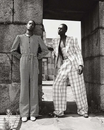 Two Black male fashion models in patterned suits pose near a stone archway, showcasing a black and white editorial fashion aesthetic.