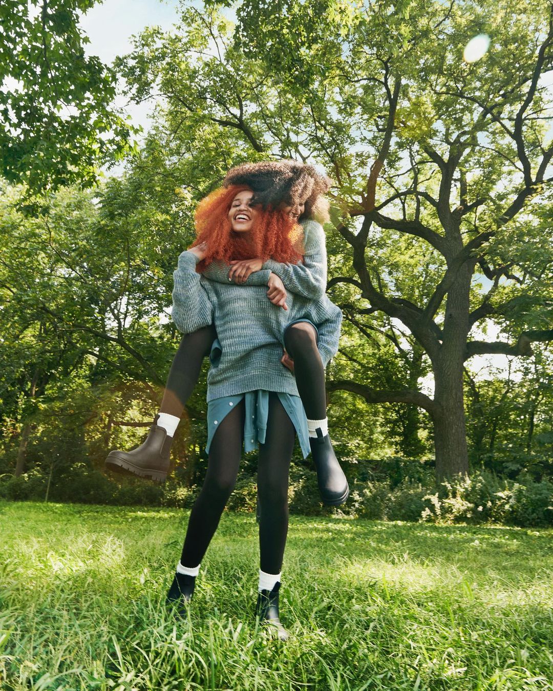 Two women in an outdoor setting; one woman is giving the other a piggyback ride, wearing sweaters, dark tights, and boots in nature.