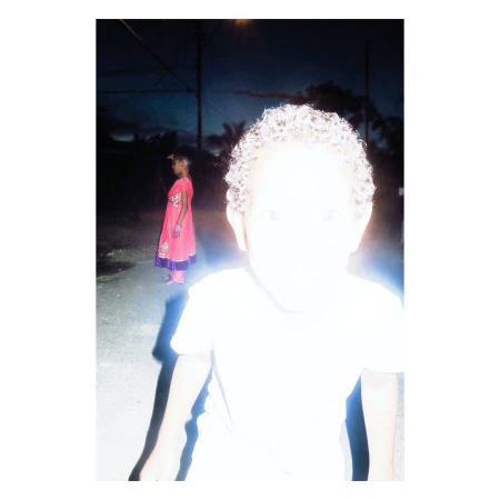 Close-up portrait of a child with curly hair completely overexposed by direct flash, with another child in the background at night