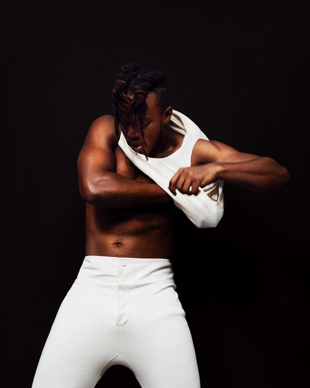 Muscular African American man with unique hairstyle removes his white tank top in a studio setting against a dark backdrop.