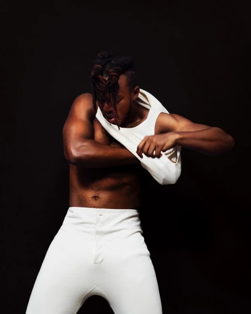 Muscular African American man with unique hairstyle removes his white tank top in a studio setting against a dark backdrop.