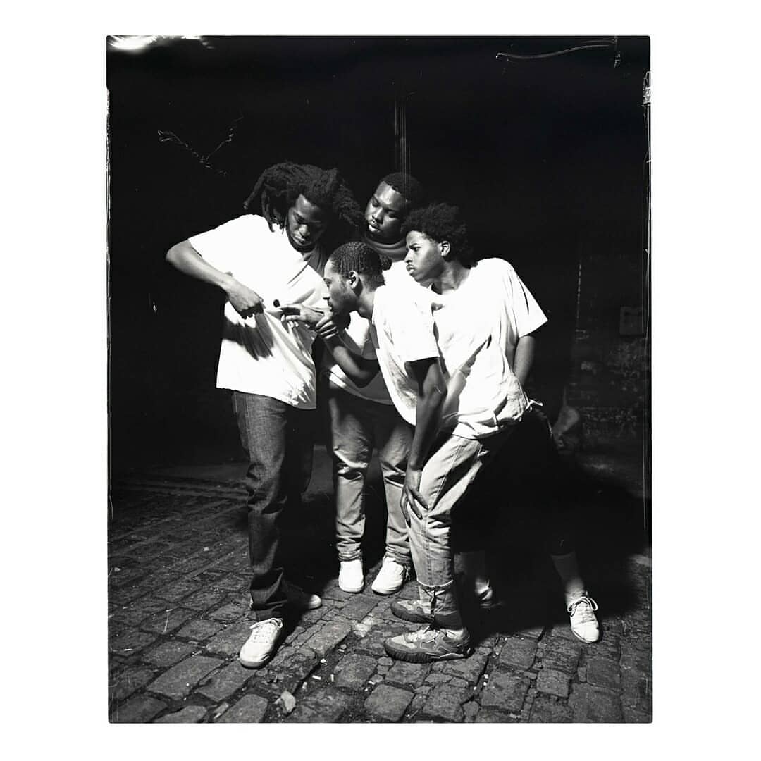 Monochrome portrait of five young adults outdoors, all looking in the same direction while standing on a brick-paved surface.