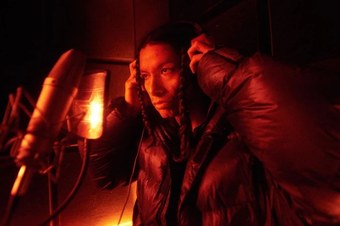 Young man with braids wearing headphones singing into microphone in a red-lit music recording studio.