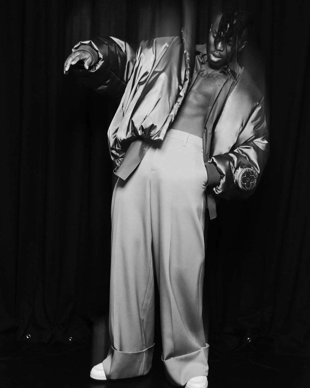A bare-chested person in light trousers and a shiny bomber jacket poses against a dark background, showcasing fashion and style.