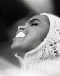 Profile portrait in black and white of a woman wearing a textured knit hood with a white object held between her teeth.