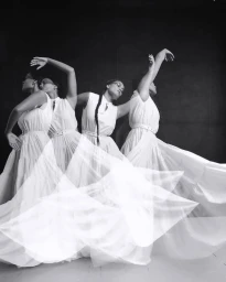 Multiple exposure black and white photograph of a woman dancing in a flowing white dress against a dark studio background.