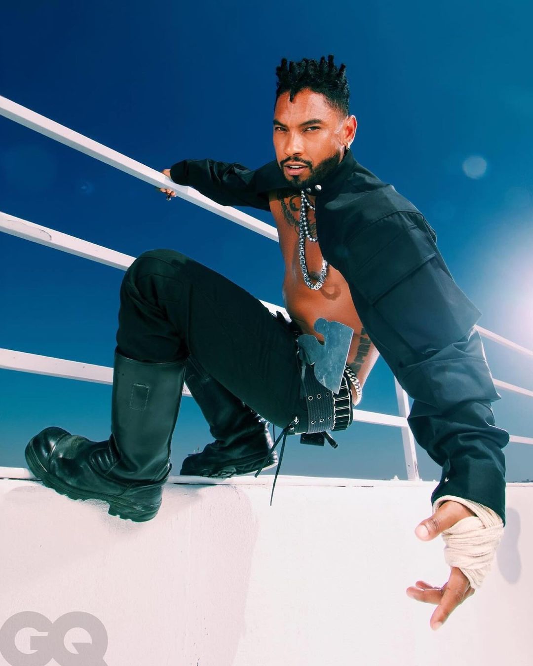 Stylish man posing in black clothing with heavy boots. He is outdoors against a blue sky and white railing on a sunny day.