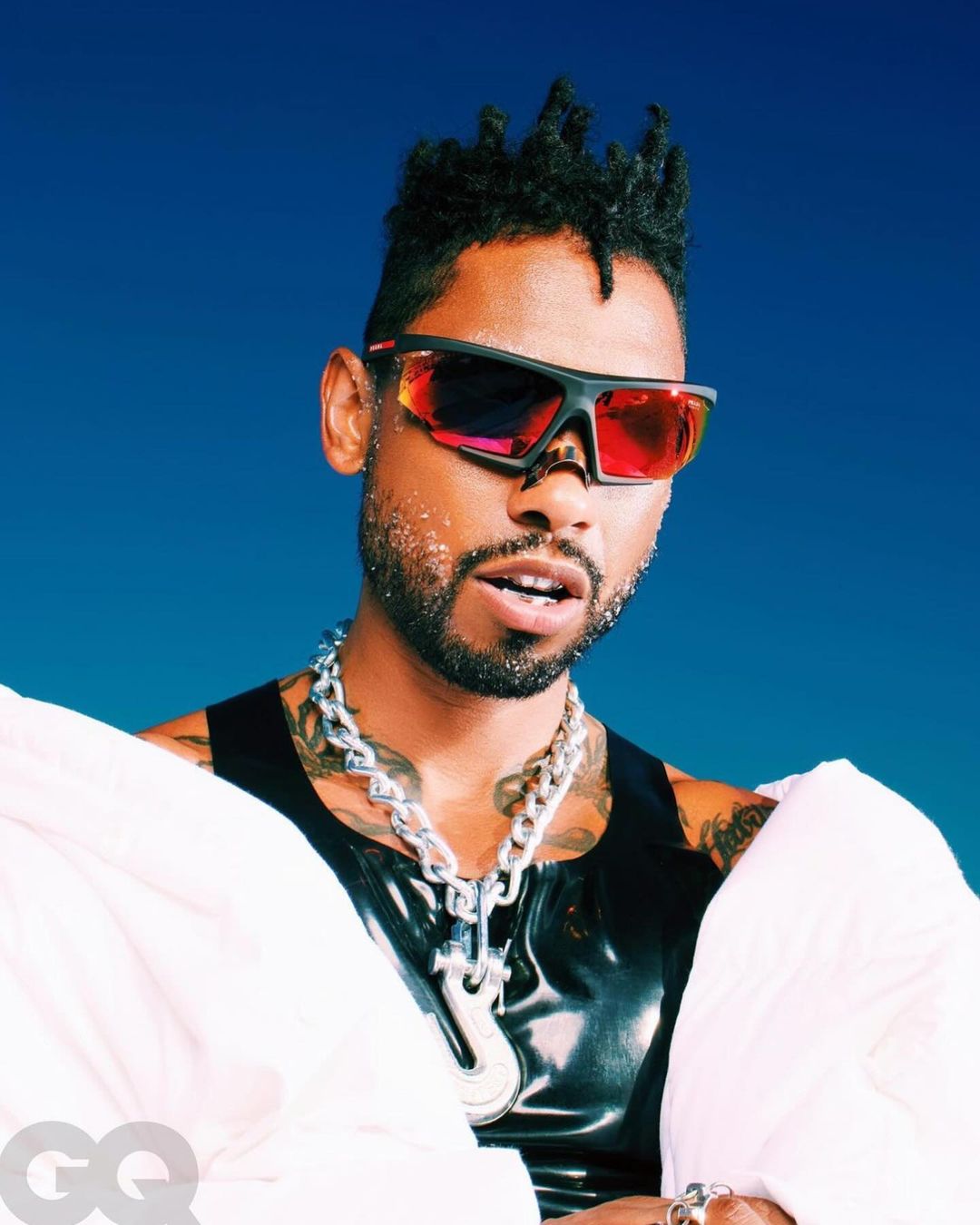 Close-up portrait of an adult male with dreadlocks and sunglasses, wearing a tank top and chain necklace in a blue studio setting.