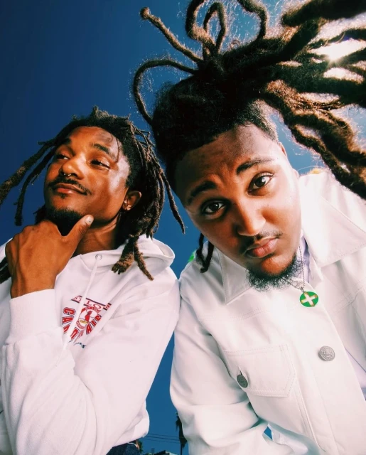 Two Black men in hoodies and a white jacket pose against a bright blue sky, captured with a wide-angle lens for a unique perspective.