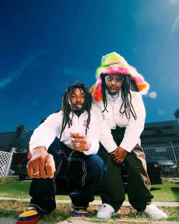 Two black men outdoors in Los Angeles, one crouching in white jacket and dark pants, the other standing wearing a fluffy multi-colored hat