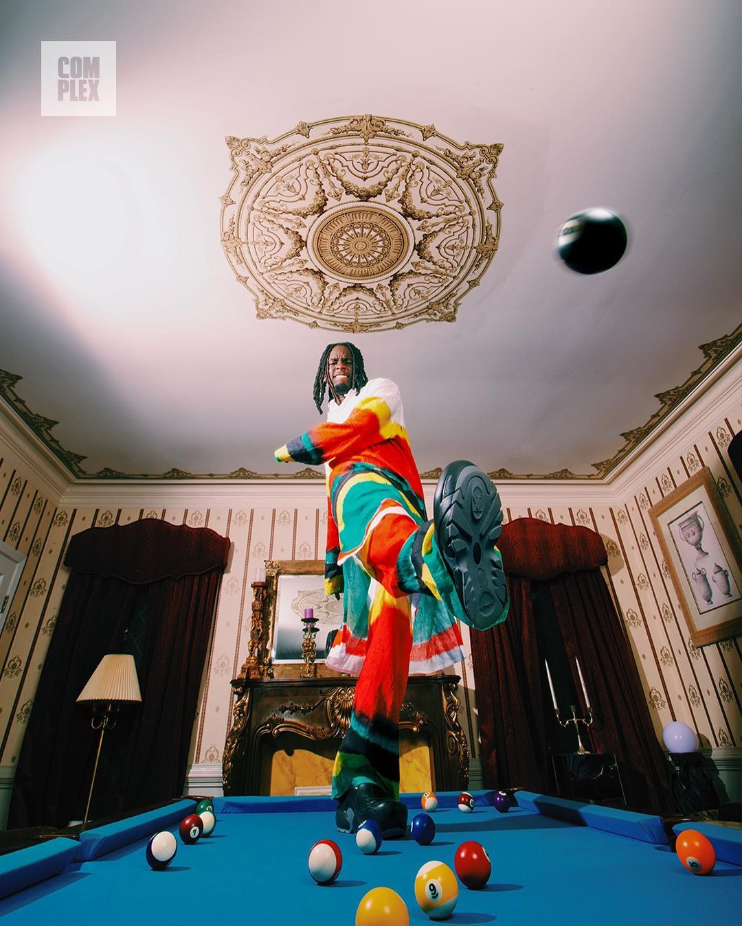 A man in colorful patterned clothing kicks a billiard ball on a pool table in a room with ornate architectural details.