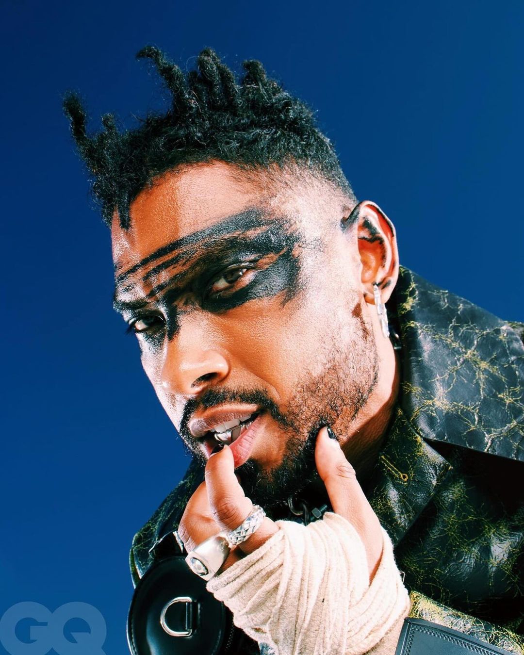 Portrait of a man with dreadlocks, black eye makeup, and a patterned jacket against a blue background, showcasing fashion and style.