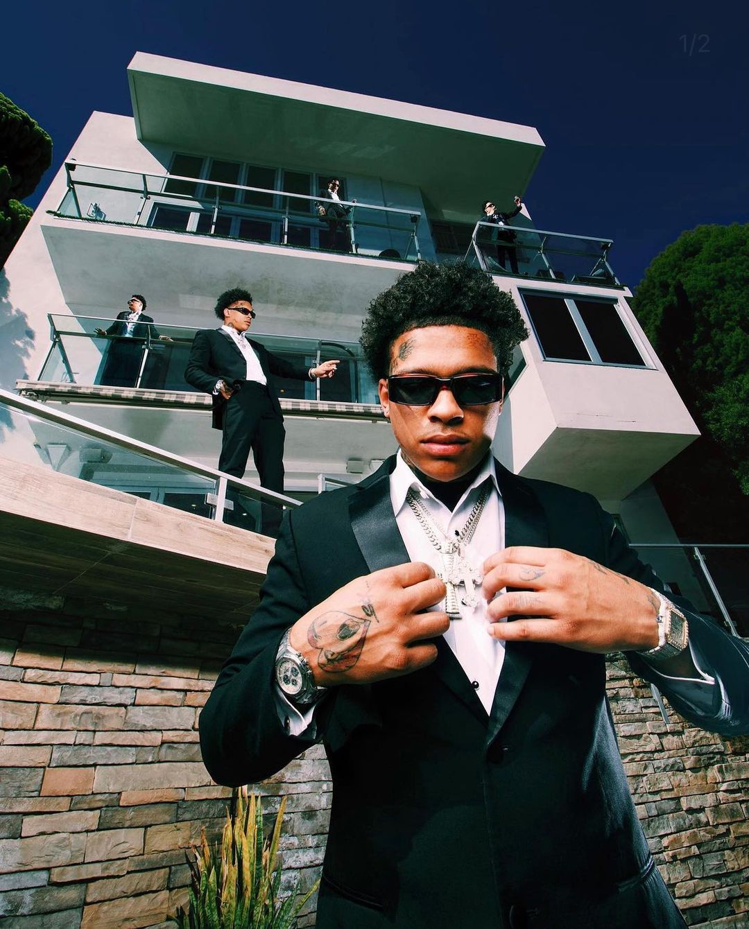 Stylish adult male in a black tuxedo and sunglasses, standing in front of a modern home with other men on the balconies.