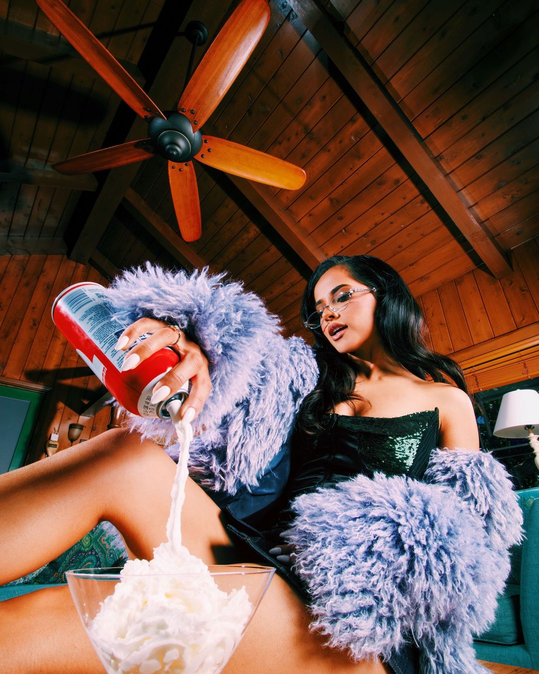 A young adult woman in a plush jacket pours whipped cream into a large glass indoors, with a wood-paneled ceiling and fan in the background.