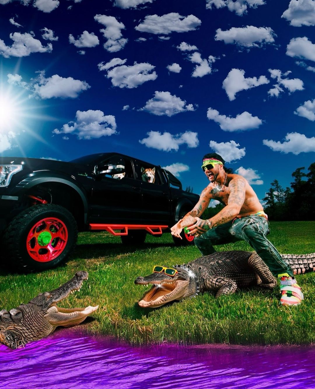 Shirtless tattooed man with sunglasses poses in a field with two alligators, next to a black truck and two dogs under a bright sky.