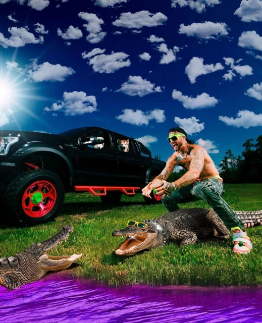 Shirtless tattooed man with sunglasses poses in a field with two alligators, next to a black truck and two dogs under a bright sky.