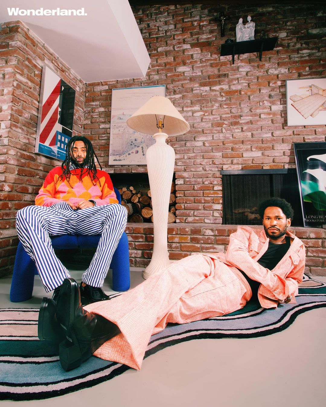 Two men pose indoors on a striped rug in stylish outfits: one seated wearing a multicolored sweater and the other reclining in a pink suit.
