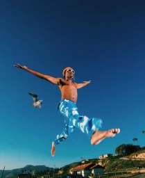 Shirtless man in blue patterned trousers leaping against a deep blue sky with a flying seagull