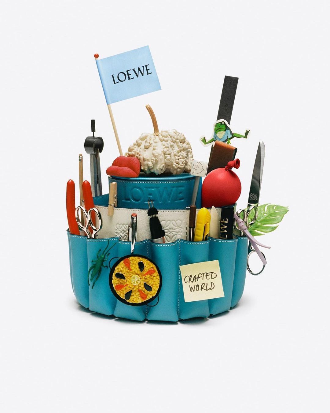 Craft supplies and various objects are arranged in a tiered blue container, with a Loewe flag and a 'Crafted World' sticky note.
